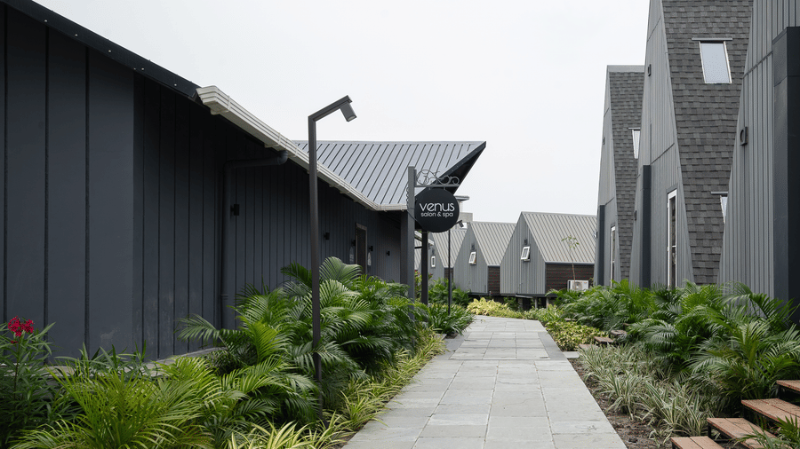 Exterior view of a paved pathway running between a row of modern, A-frame style accommodations and planters with foliage at Hotel Sonar Bangla, Taki.
