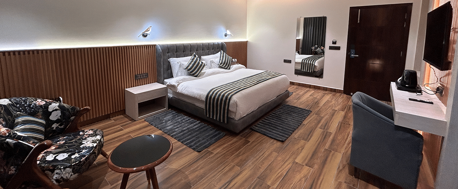 A shot of the deluxe room featuring a bed, bed side tables, two chairs, coffee table, work desk, a couch, a flat screen TV, and a full length mirror - DLS The Emerald, Nainital