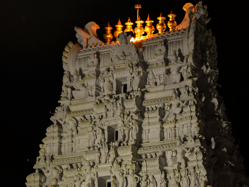 A tall, intricately carved white stone temple gopuram lit up at night with golden ornaments at the very top against a black sky.
