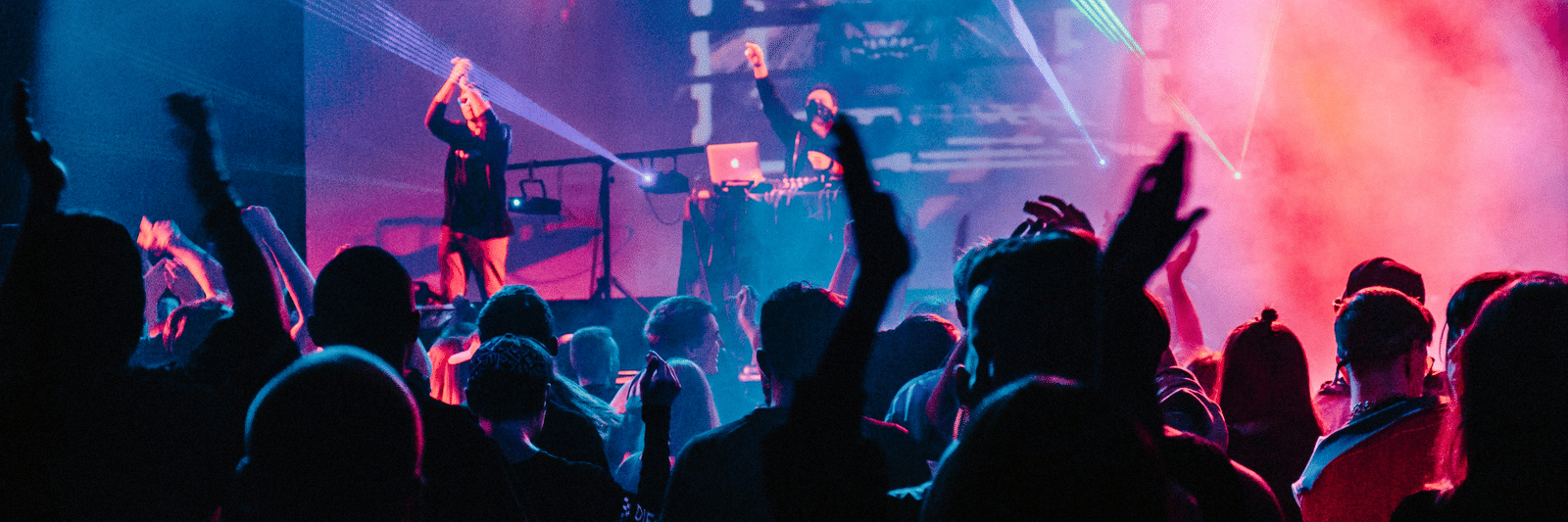Silhouetted figures of a crowd with their hands raised during a live music performance with purple and blue lighting.