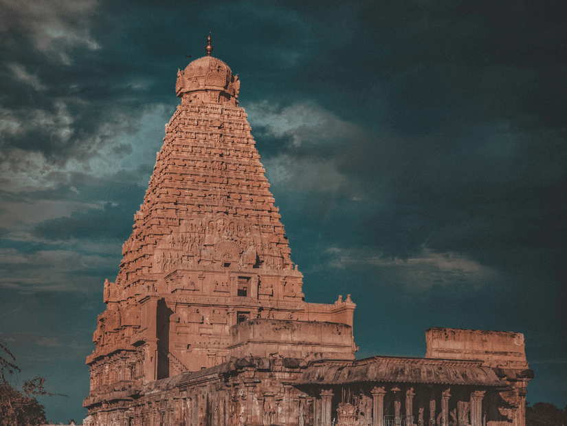 A facade view of Brihadeeswarar Temple in Thanjavur in the evening time with a person sitting in front of the temple. The climate in Thanjavur is very tropical and humid throughout the year.