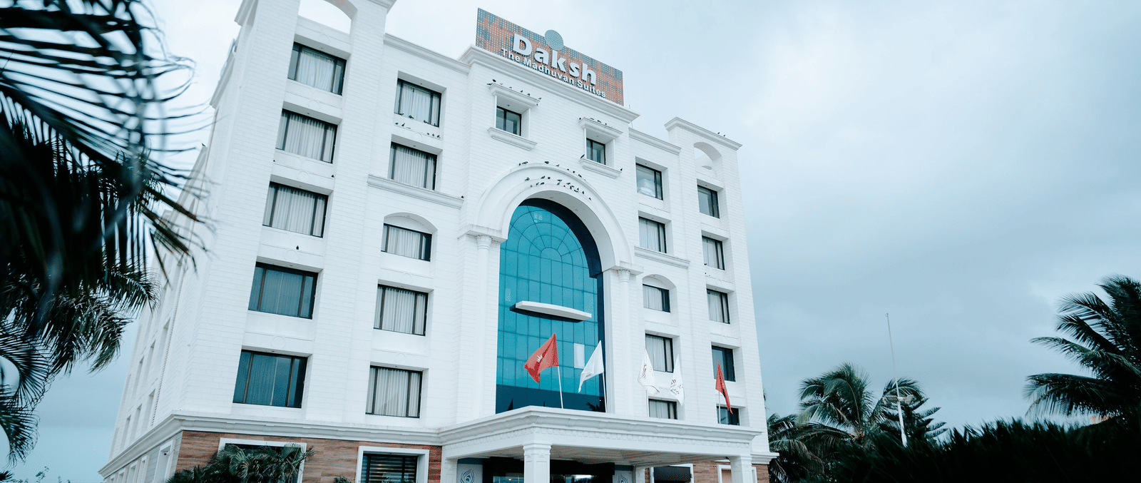 Large, modern white hotel building exterior on a slightly overcast day. The main entrance features a large arched blue glass window at Daksh The Madhuvan Suites, Dwarka.