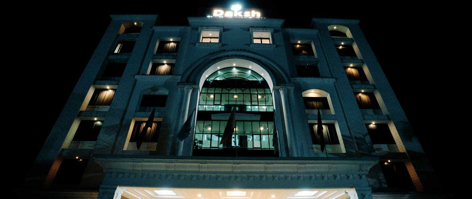 Dramatic low-angle night-time shot of the hotel's brightly lit white exterior. The arched central window and entrance are prominently illuminated at Daksh The Madhuvan Suites, Dwarka.