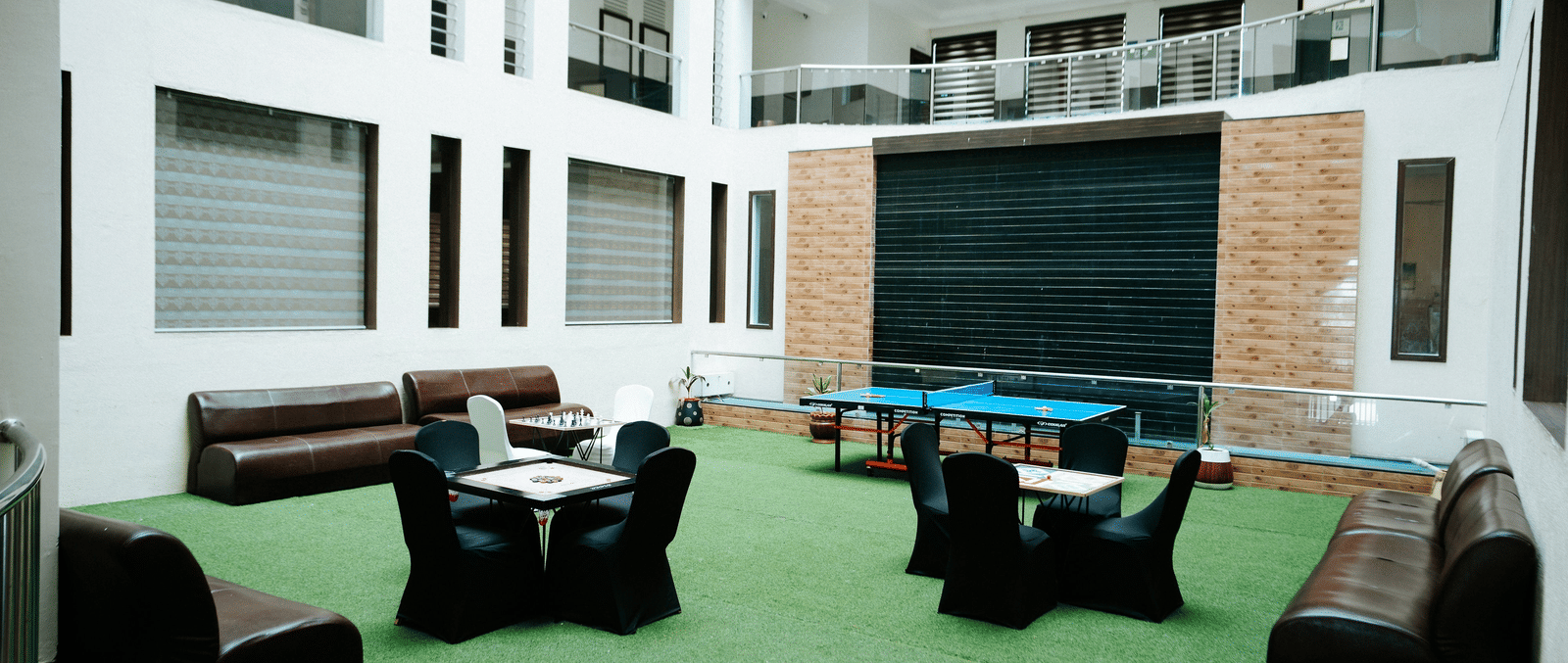 Spacious, brightly lit interior courtyard or lounge area. Features modern white walls, a green carpeted floor, and dark brown leather seating at Daksh The Madhuvan Suites, Dwarka.