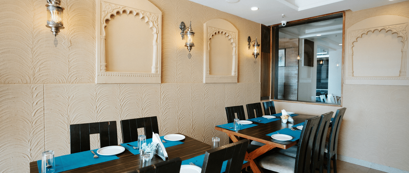 Restaurant area with tables set with blue runners, decorative beige wall arches, and traditional style wall lighting at Daksh The Madhuvan Suites, Dwarka.