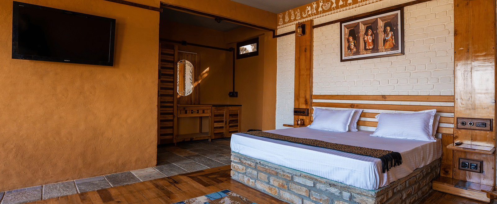 Rustic hotel room with wooden bed, wall art, TV, and patterned rug on hardwood floor. - Blusalzz Homes The Himalayan Bungalow, Almora