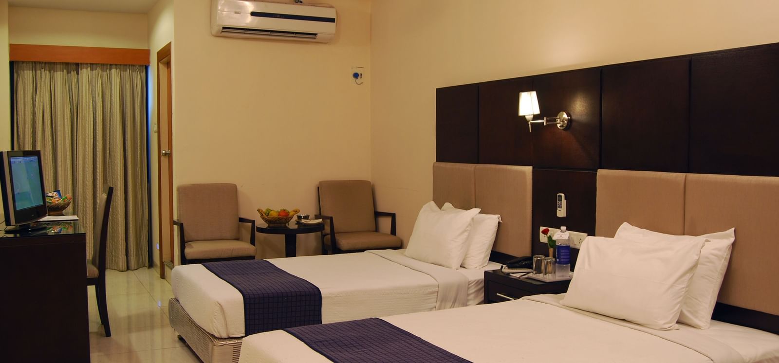 A well furnished A.C. twin room at Lords Eco Inn Dahej, featuring modern furnishings, a standalone T.V., wooden headboard, a headboard light and beige curtains on floor-to-ceiling window.