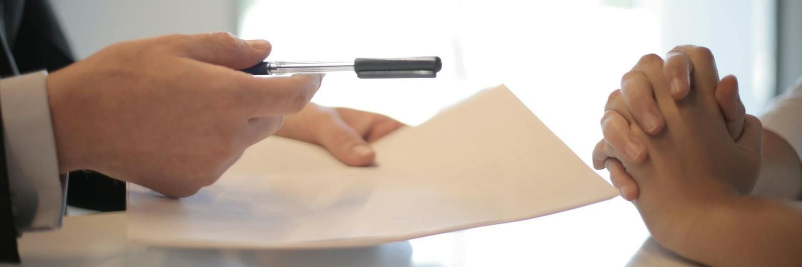 Close-up of two people's hands on a desk, one holding a pen and paper, while the other's hands is crossed, suggesting a discussion