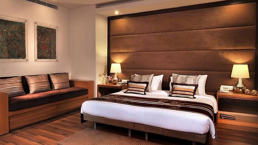 A well-lit room featuring a plush bed with lamps on both sides on the nightstands and a sofa above which 2 paintings are hanging from the wall