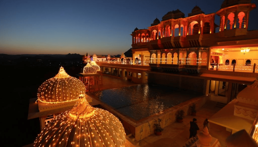 The swimming pool and terrace area on 5th floor of Chunda Palace, Udaipur, featuring traditional architecture with grand domes illuminated by fairy lights against a deep twilight sky.