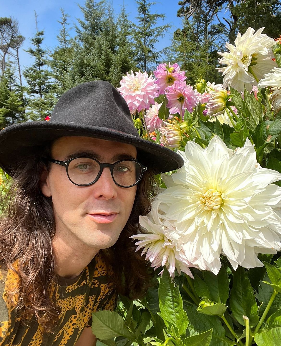 Man in glasses and hat holding a large white dahlia in a garden.