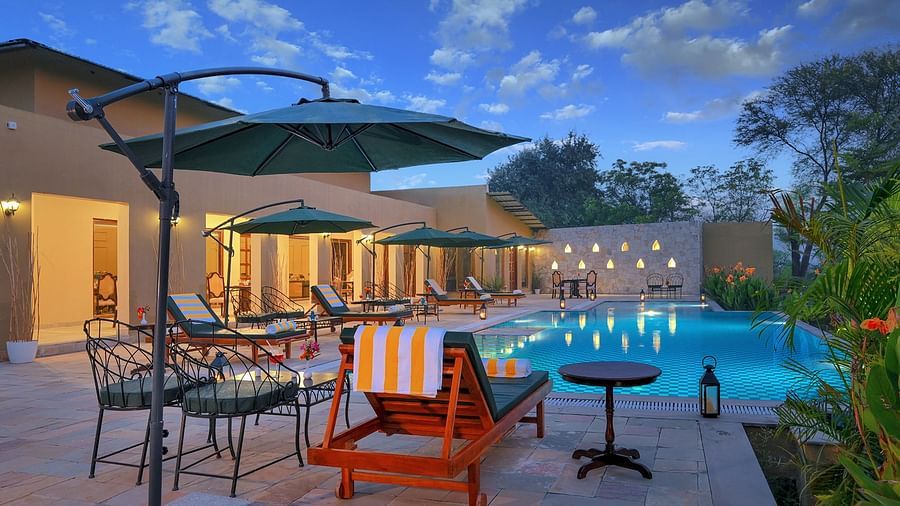 A poolside area with wooden loungers, green umbrellas, lanterns, and a stone accent wall. The pool reflects the blue sky, and archways lead to an indoor dining space with classic wooden furniture - Singhvilas, Ranthambore
