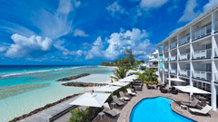 The view of The Soco Hotel facade with a bright blue pool, lounge chairs and palm trees along the coastline under a cloudy blue sky.