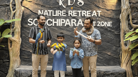 A family enjoying time with colourful parrots at RD’s Nature Retreat Soochipara Bird Zone, surrounded by lush greenery and a natural stone backdrop.
