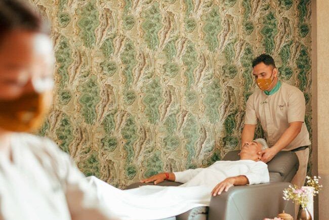 A person receiving a massage in a spa room with patterned green wallpaper and a therapist in the background at Karma Chalets.