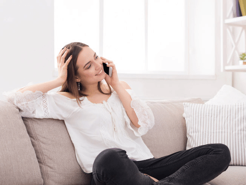 A woman sits on a couch talking on a cell phone while smiling with her hand on her head resting on the couch
