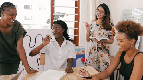 4 women engage in a lively and creative discussion around a table, surrounded by notebooks, artwork, and natural sunlight in a bright room.