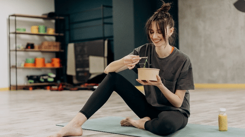 A woman sits barefoot on a yoga mat in a fitness studio, smiling while enjoying a healthy meal with a juice bottle beside her.