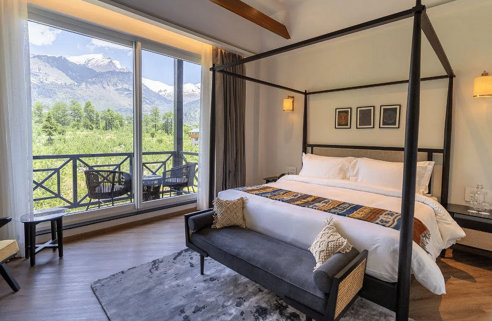 A room at Nivasana Manali featuring a four-poster bed, a couch, and a balcony with seating with stunning views of snow-capped mountains and lush greenery in the foreground.