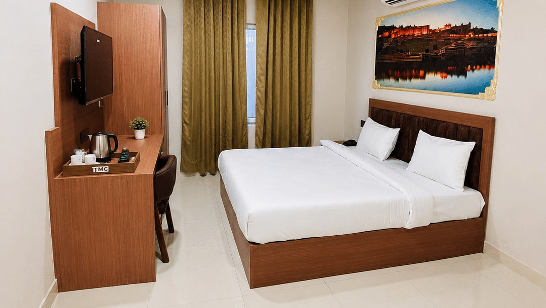 A bedroom inside Lime Tree Hotel & Banquet, Jaipur. The room contains a bed, a television on a desk, a window covered by curtains, and a door.