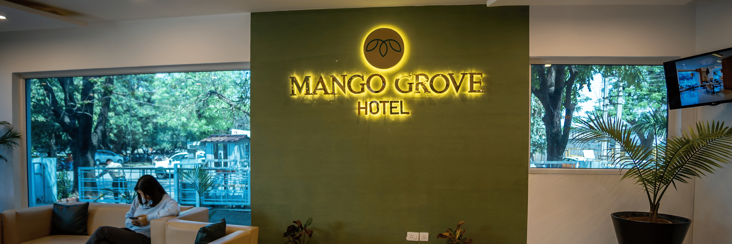 Lobby seating area with a sofa and the hotel logo displayed on a green accent wall at Mango Grove Hotel.