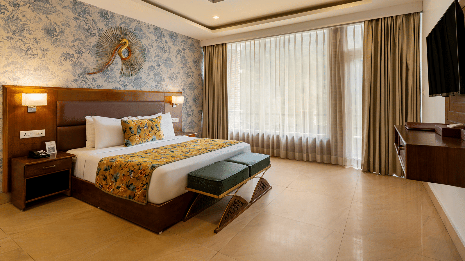 A spacious bedroom featuring a neatly made bed, 2 side cabinets, a wall with blue wallpaper, a decorative ceiling, and sheer and opaque curtains | Summit by the Ganges Beach Resort & Spa, Rishikesh