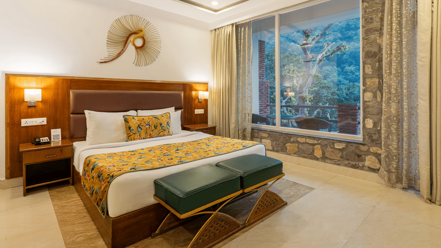 A spacious bedroom with attached balcony, featuring a neatly made bed, 2 vanity stools with leather cushions, 2 bedside tables, and a view of the trees outside | Summit by the Ganges Beach Resort & Spa, Rishikesh