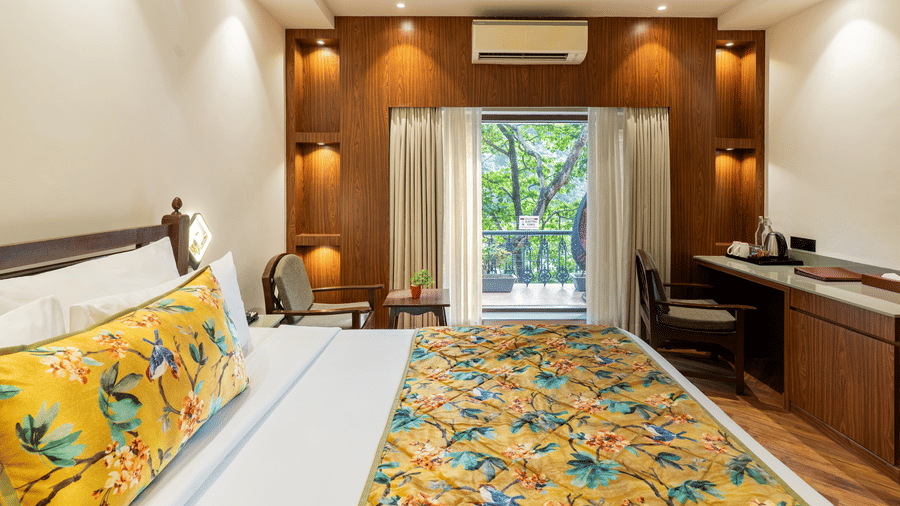A bedroom featuring a big bed, 2 wooden chairs, an A/C unit, an attached balcony with a standing swing chair, potted plants, and view of the trees outside | Summit by the Ganges Beach Resort & Spa, Rishikesha
