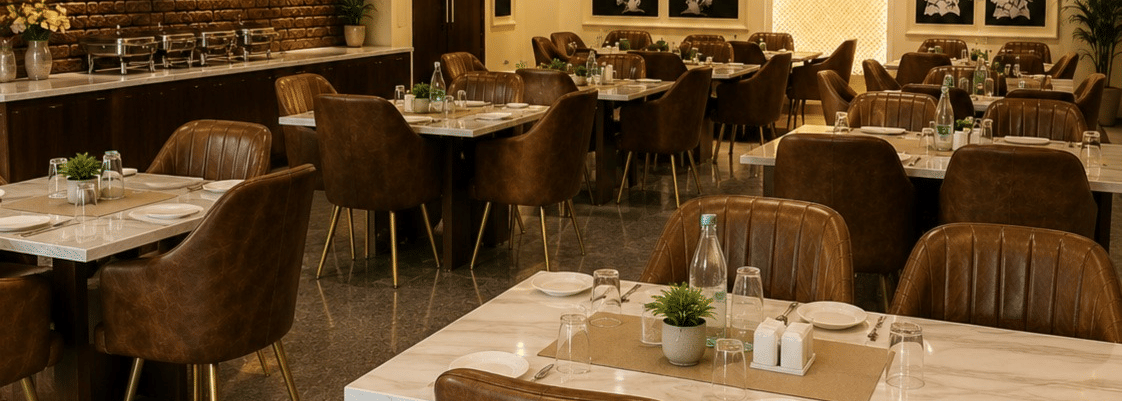 An indoor dining area or restaurant space with several set dining tables and brown chairs arranged under warm ceiling spotlights.