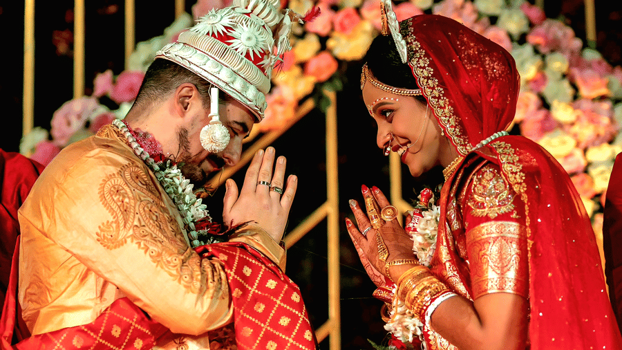 A bride and groom in traditional wedding attire sharing a ceremonial moment under a colorful floral arch at Vedic Village Spa Resort with pink and white flower decorations surrounding them.