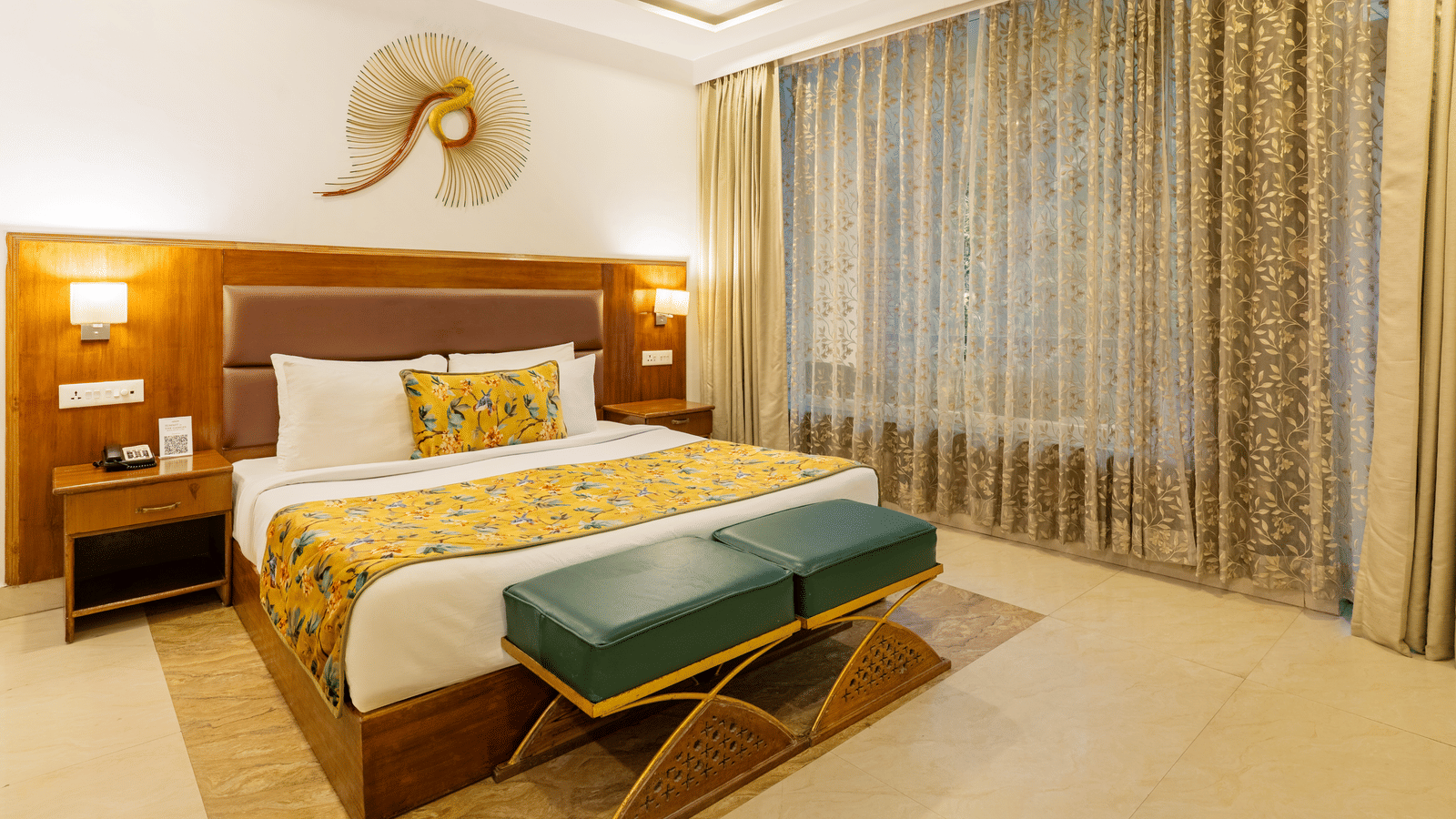 A room featuring a wooden bed, 2 bedside tables, 2 vanity stools, a decorative wall piece, and full-length curtains | Summit by the Ganges Beach Resort & Spa, Rishikesh