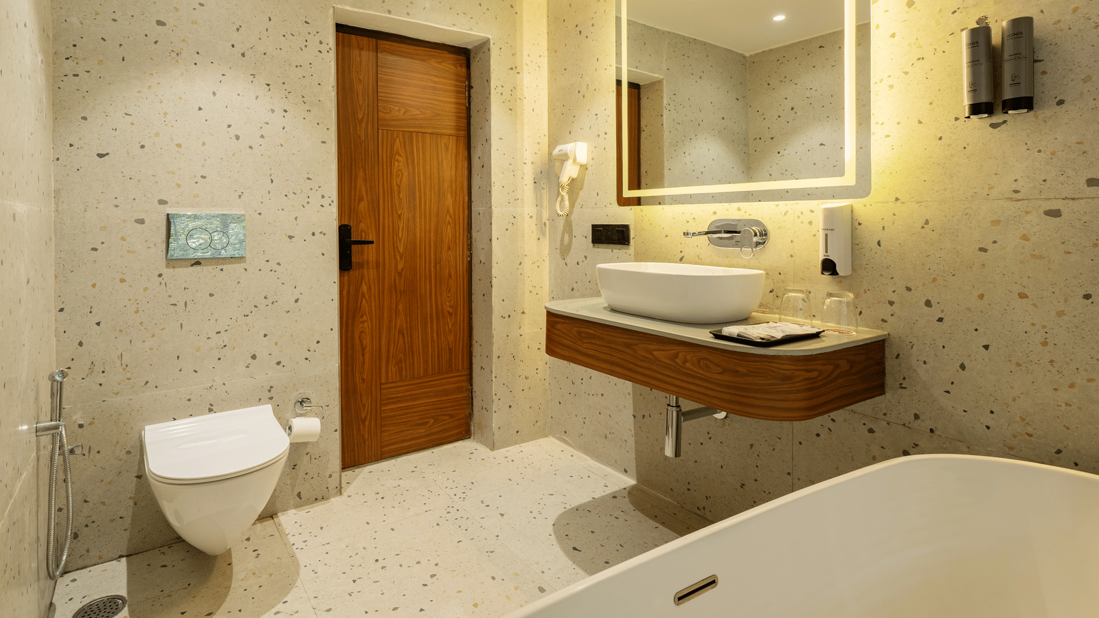 A bathroom featuring a big mirror, a wash basin on a counter with toiletries, soap dispensers and a hair dryer on a wall, and a closed door | Summit by the Ganges Beach Resort & Spa, Rishikesh