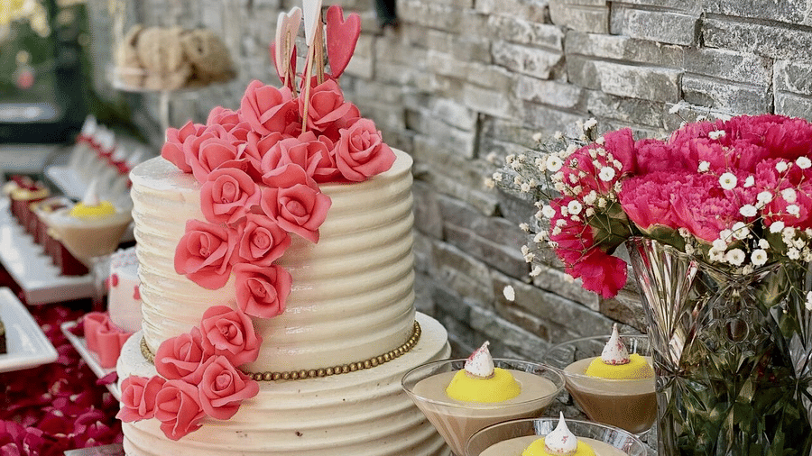 Elegant dessert table featuring a two-tier cream cake with pink sugar roses, pastries, mousse glasses, and fresh flowers.