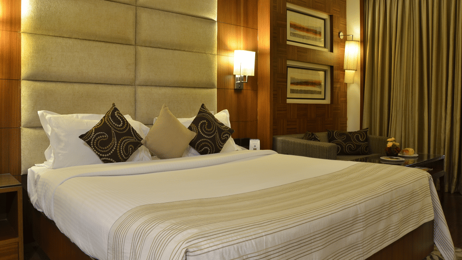 Premium Room at The Bristol Hotel, Gurgaon with a king-size bed, tall padded headboard, brown patterned cushions, framed wall art, wall sconces, and a sofa.