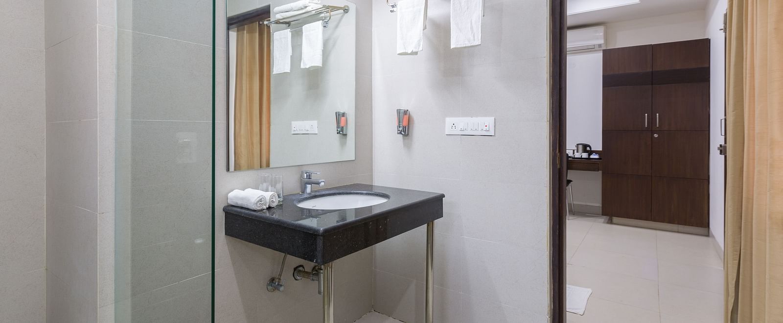 A bathroom with a dark sink, mirror, and a glimpse into the bedroom - Sanctum Suites BEL Road, Bangalore