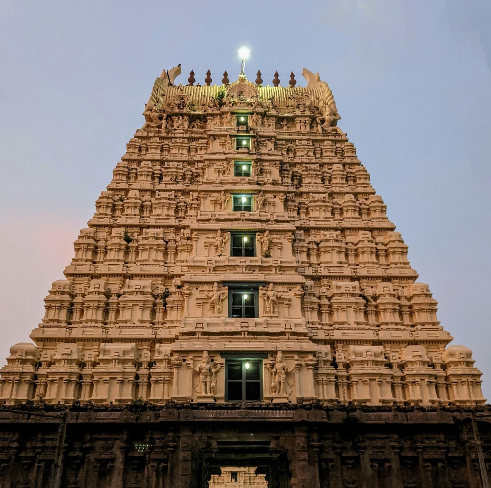 A majestic view of the Tirupati Balaji Temple gopuram, illuminated against a twilight sky. The intricate stone carvings and towering structure are prominent.