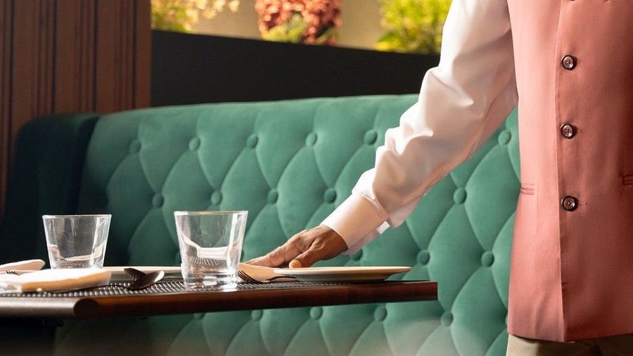 A staff member at Clarks Inn, Basti, wearing a shirt and a vest, sets a table in a restaurant with tufted seating.
