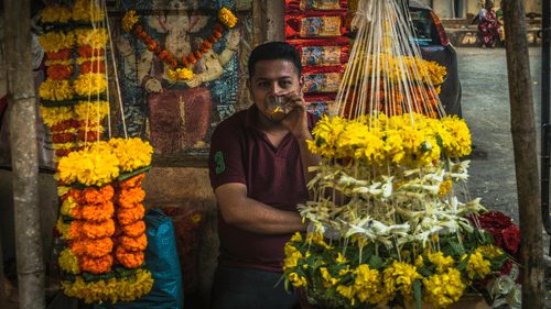 A man drinking beverage standing behind garlands with a painting of lord Ganesha on the wall in the background
