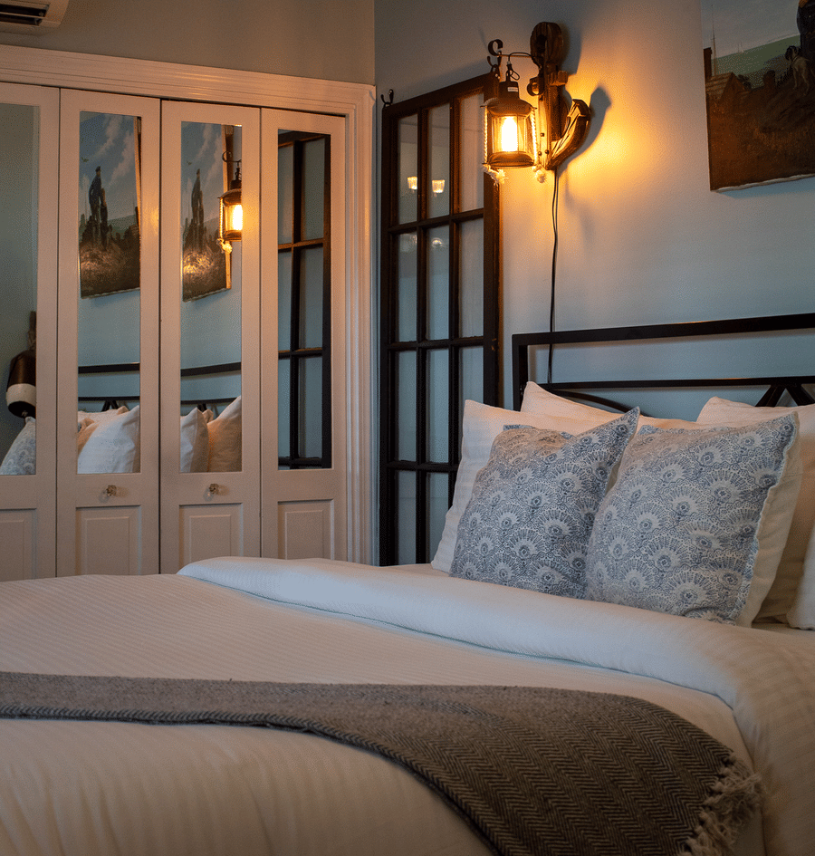 A close-up view of a bed with white bedding, blue and white pillows, and a gray throw blanket, with a black metal headboard and a wall-mounted light fixture.
