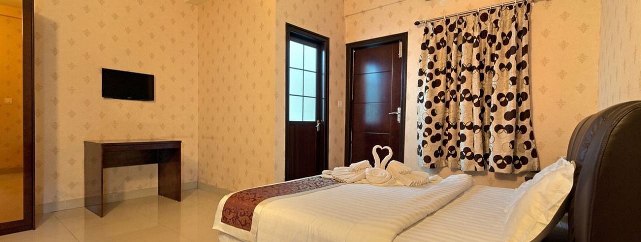 Another view of a hotel room at TGI Livancia Hillside, JC Nagar, Mysore, showing a bed with towel swans, a television on the wall, and a glimpse of the room's decor.