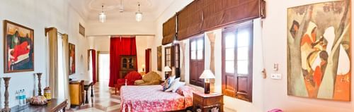 A suite at Neemrana's - Hill Fort - Kesroli features beds with covers, bedside tables with lamps, seating with a couch and chairs, tables, artwork, windows with blinds, and tiled flooring.