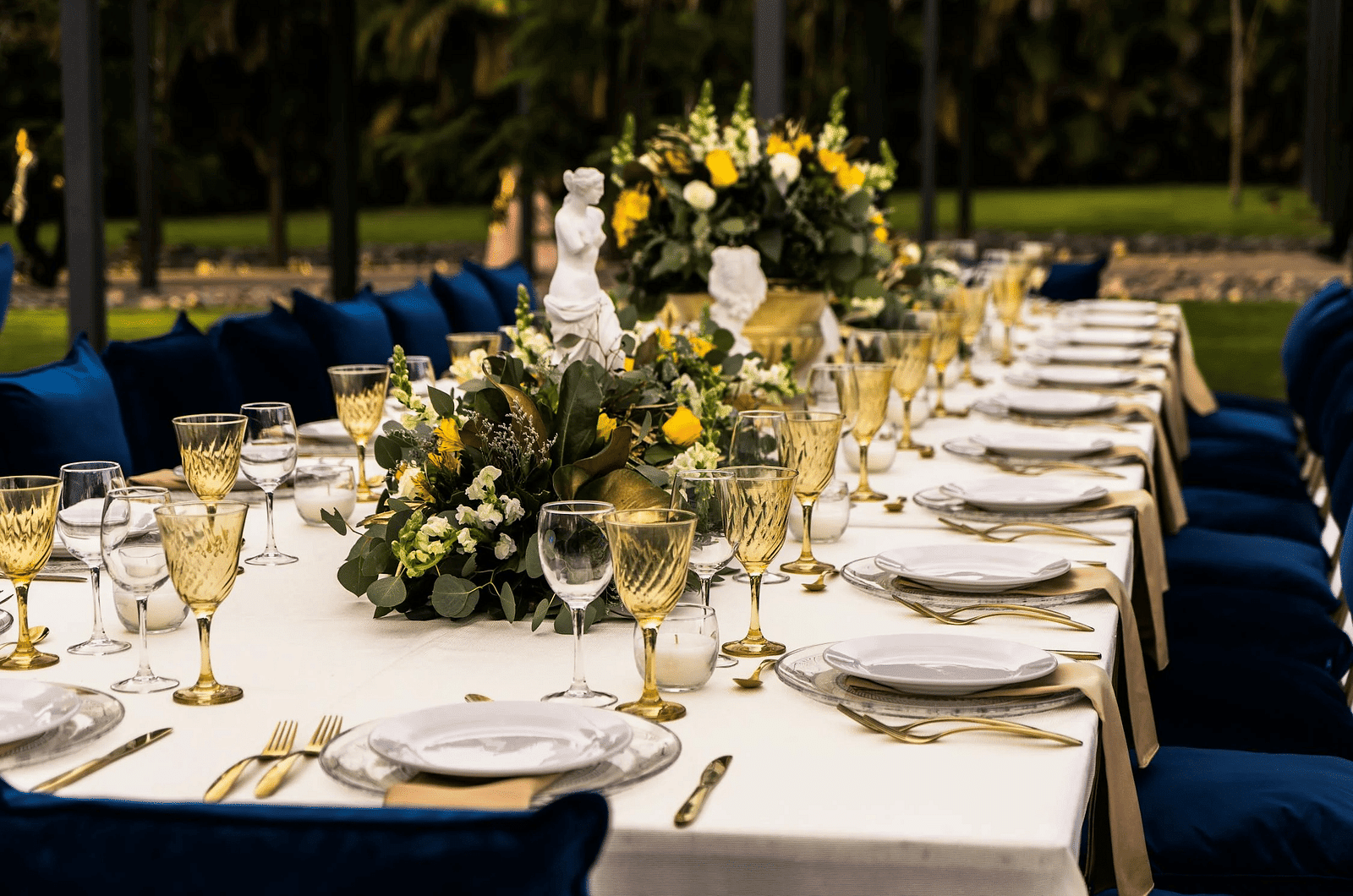 A rectangular table in an outdoor setting with cutleries arranged on it.