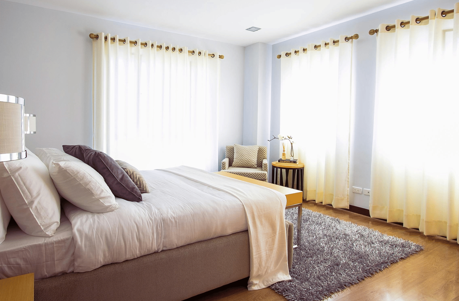 A neatly arranged bedroom with a large bed, pillows, side tables, armchair, and soft natural light filtering through curtains.