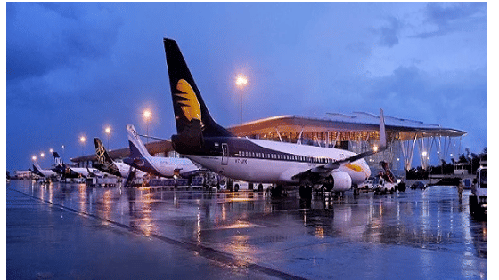 A view of the wet tarmac at Bengaluru Airport at night, featuring several parked airplanes, including a Jet Airways aircraft, reflecting bright airfield lights on the rainy ground.