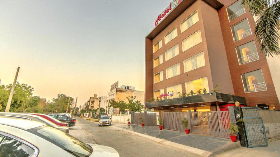 A wide-angle street view of the modern hotel building facade with cars parked along the road at sunset - Hotel 91 HUDA City Centre, Gurugram