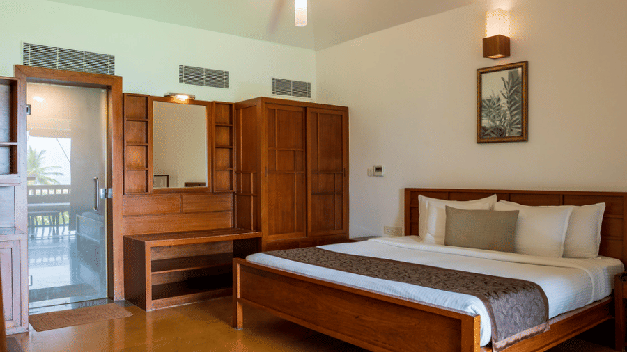 A hotel room featuring a double bed, a wooden wardrobe with a vanity mirror, and a glass door leading outside | Le Pondy