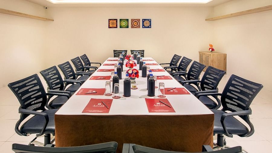 A formal meeting room is set up with a long rectangular table surrounded by numerous black office chairs, featuring water bottles and red and white placemats, under bright ceiling lights and with framed artwork on the wall - Lords Inn Motikhavdi, Jamnagar.