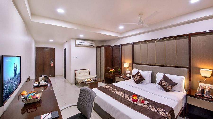 A well-lit hotel room offers a king size bed with decorative pillows, a work desk with a television, and a glimpse into the bathroom.