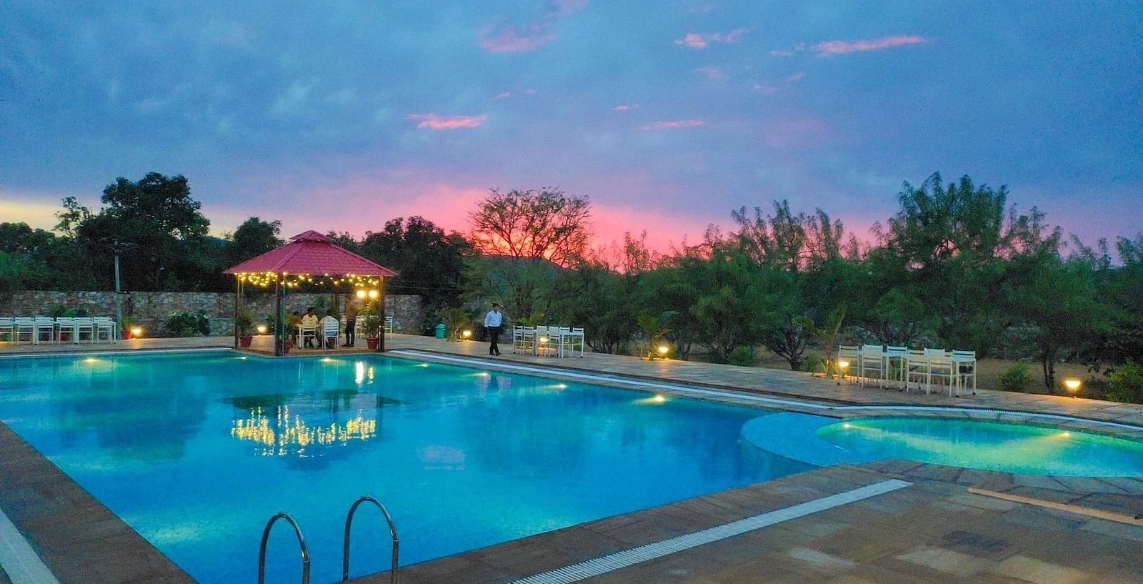 An outdoor swimming pool at dusk with a pink and blue sky in the background - Bamboo Saa Mulberry Resort, Pushkar