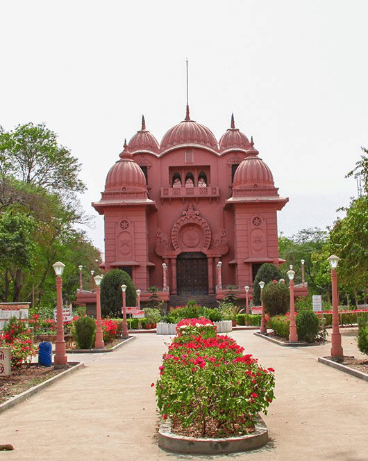The symmetrical temple at Ramakrishna Ashrama, Rajkot, featuring multiple domes and a carved entrance, with a path lined with flowerbeds and lamp posts.